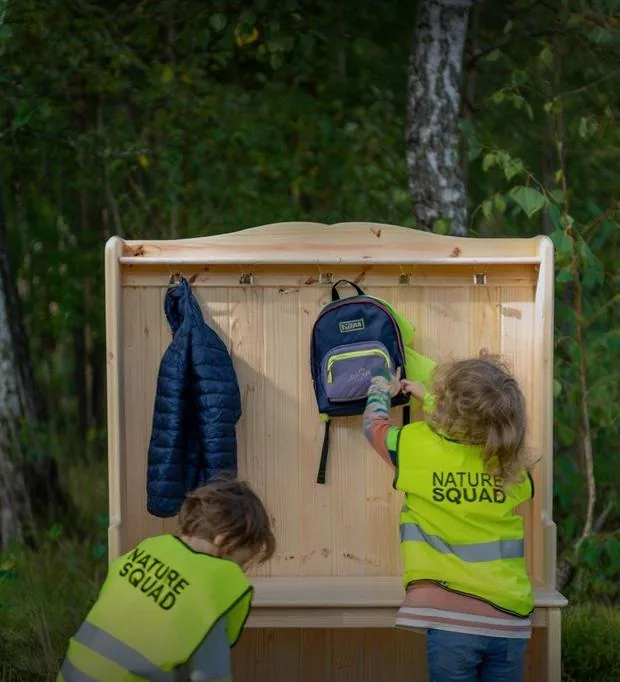 Due bambini in gilet Nature Squad appendono zaini e giacche alla panca di legno nel bosco